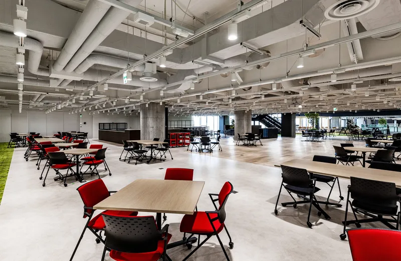 Open office floor with exposed ceiling ductwork and red chairs arranged around light wood tables