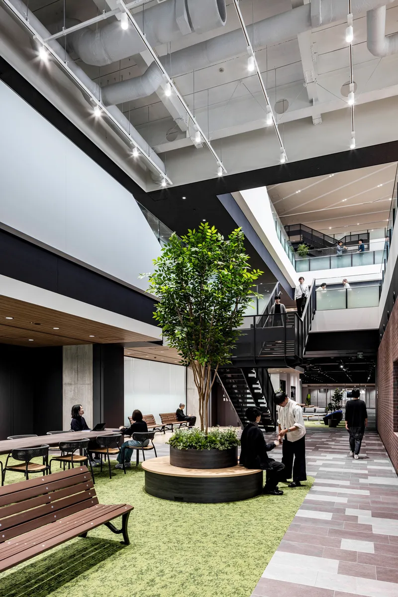 Multi-level atrium with central tree planter, green turf seating area, and black steel staircase connecting upper floors