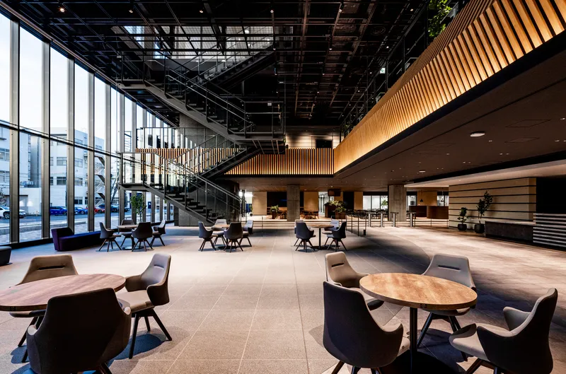 Ground floor lobby with floor-to-ceiling glazing, timber-clad mezzanine overhang, and scattered café tables
