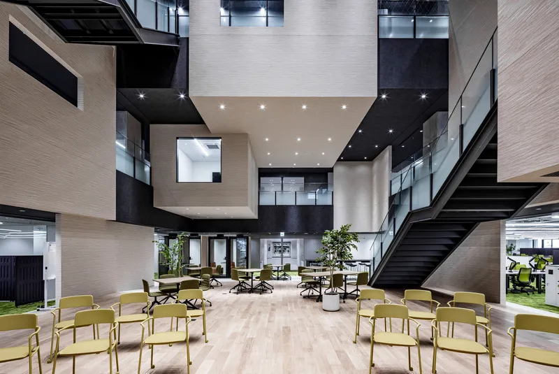 Multi-storey atrium with light wood-clad volumes, black ceilings and yellow chairs arranged on the ground floor