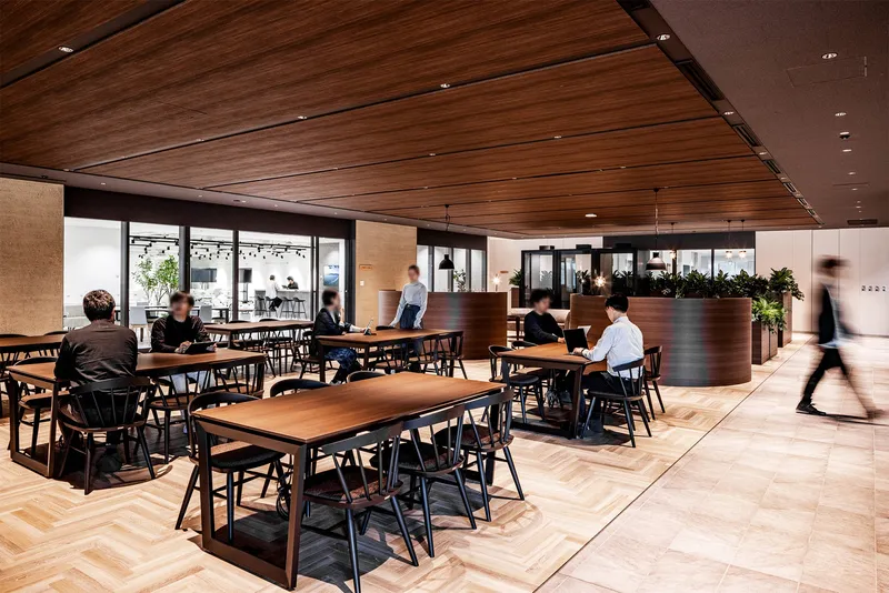 Open dining area with wooden tables and black chairs beneath a slatted timber ceiling with people in motion