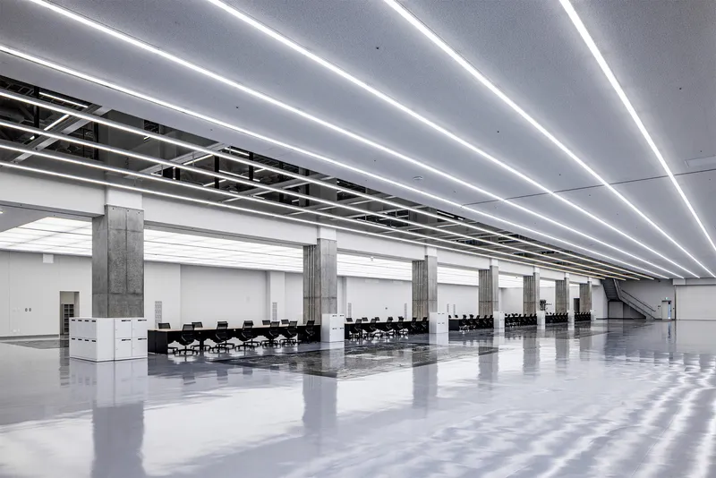 Long hall with polished floor, concrete columns and parallel linear lighting strips receding toward conference tables