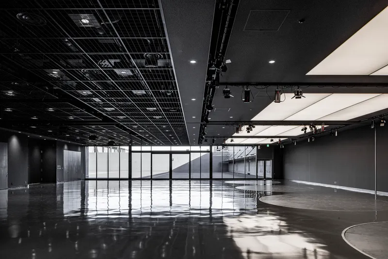 Open event space with black exposed ceiling grid and glazed walls overlooking an interior staircase
