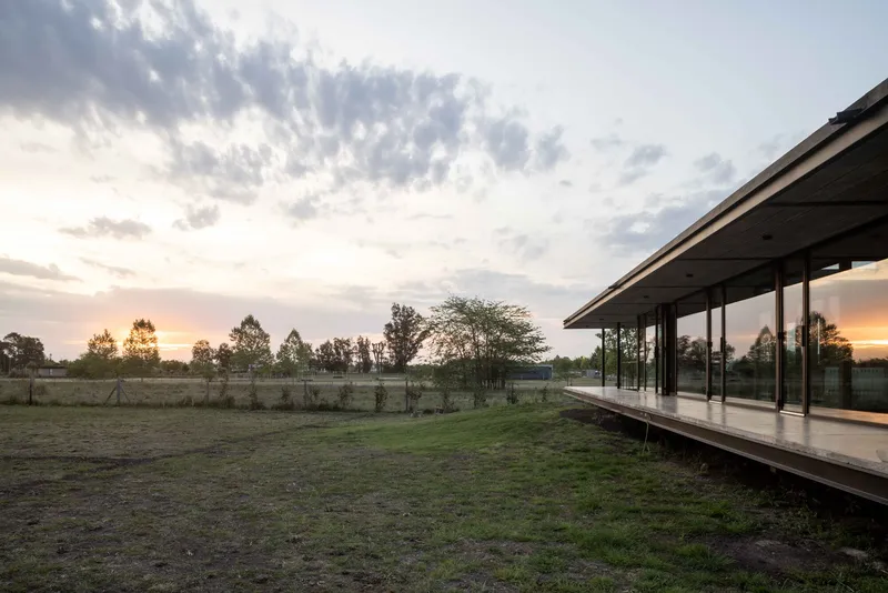 Elevated glass pavilion on a raised platform overlooking open grassland at sunset