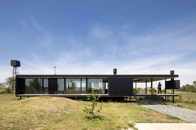 Elevated black corrugated metal pavilion with full-height glazing and covered terrace in open grassland