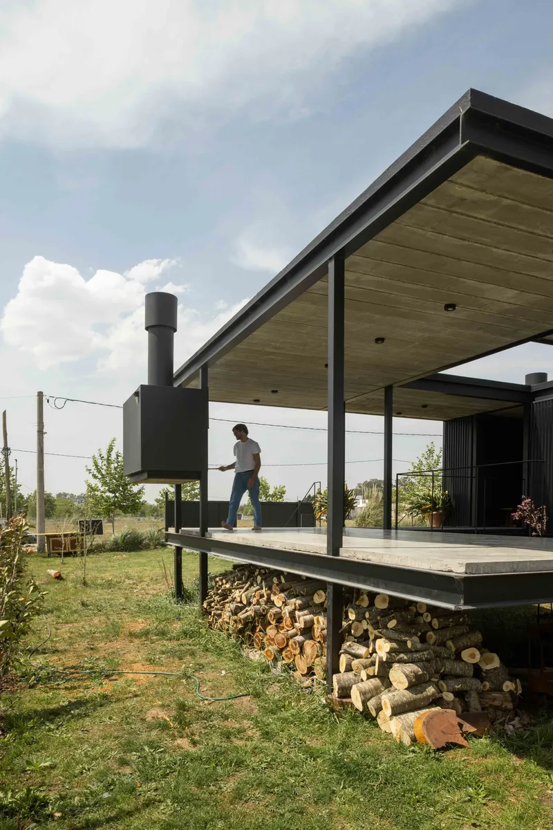 Covered deck with stacked firewood storage underneath and a figure walking under cloudy skies
