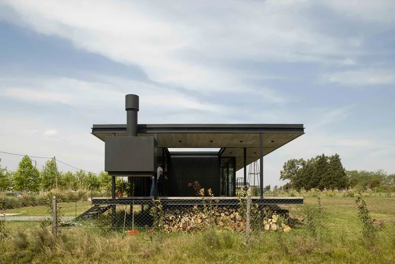 Single-story pavilion with deep roof overhang and exterior fireplace chimney in an open grassland