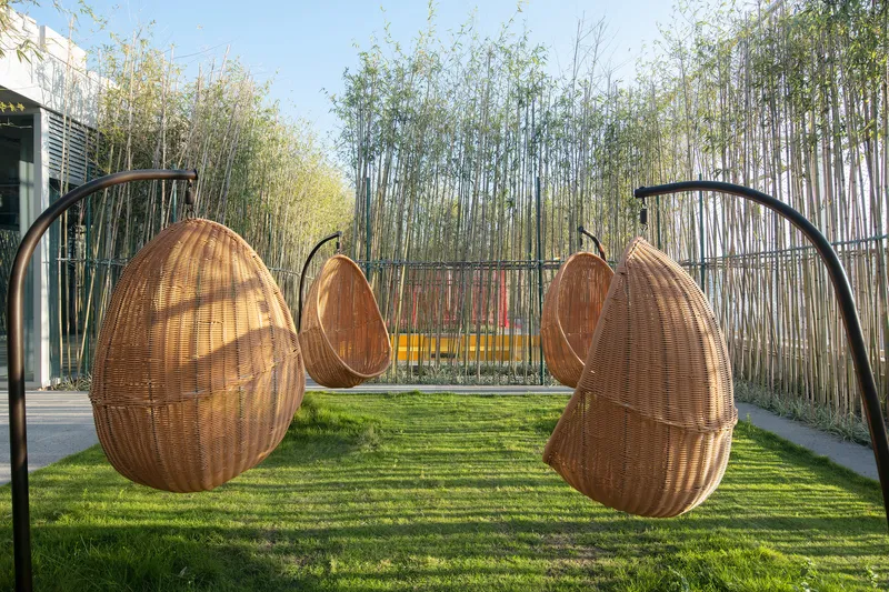 Woven rattan hanging egg chairs suspended from metal arcs over grass with bamboo fence beyond