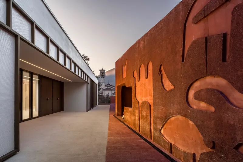Courtyard passage between weathered steel sculptural wall and white volume with vertical glazing at dusk