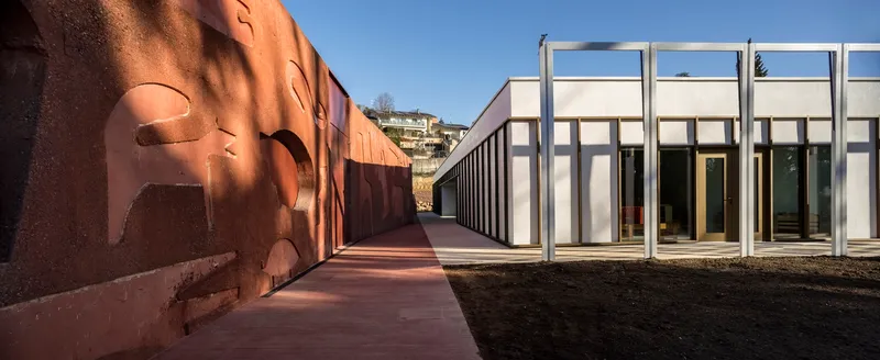 Exterior passageway with terracotta paving running between corten steel wall and white framed colonnade