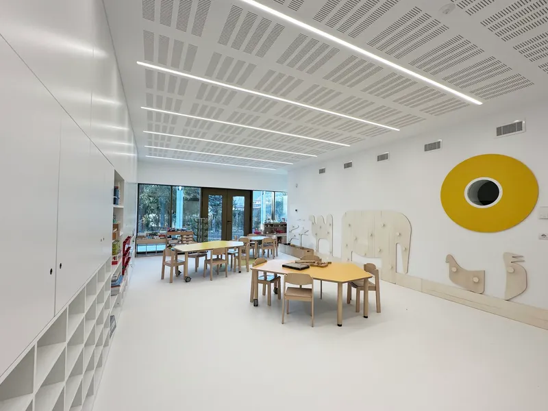 Classroom with plywood tables and chairs under perforated ceiling with linear luminaires and yellow circular wall element