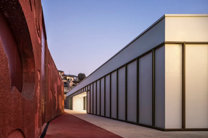 Narrow courtyard between the rammed earth wall and the translucent panel facade at dusk