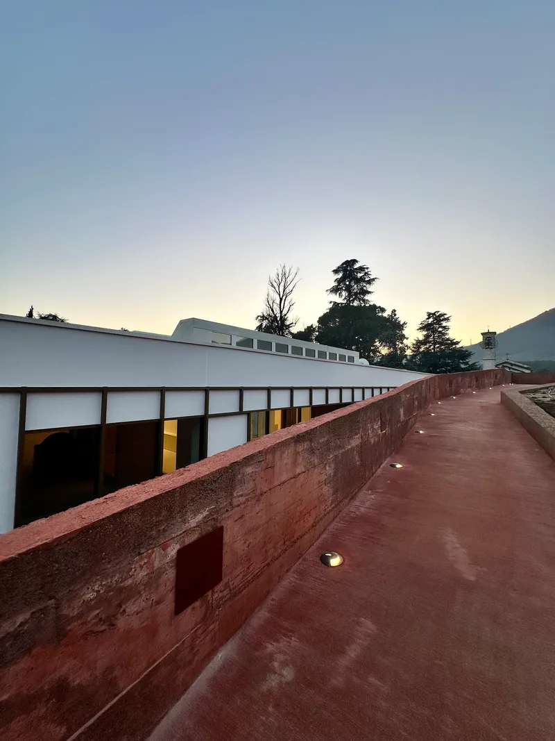 Rammed earth ramp ascending alongside the glazed facade under a pale twilight sky