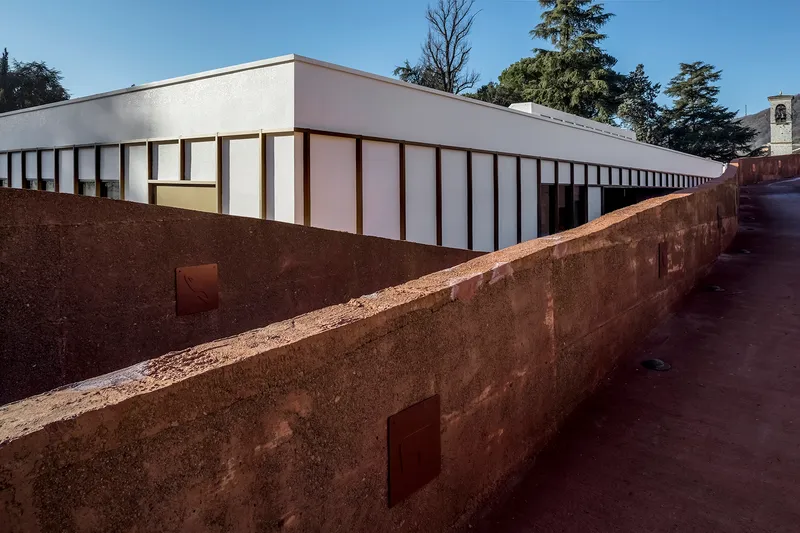 Long view of the white panel facade above the weathered rammed earth wall in daylight