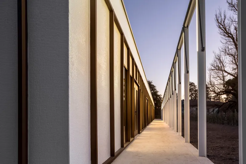 Covered walkway between textured panel walls with bare trees visible through the open side at sunset
