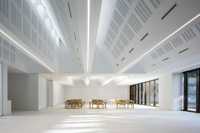 Interior hall with angled ceiling beams, linear lighting slots, and perforated acoustic panels above wooden tables