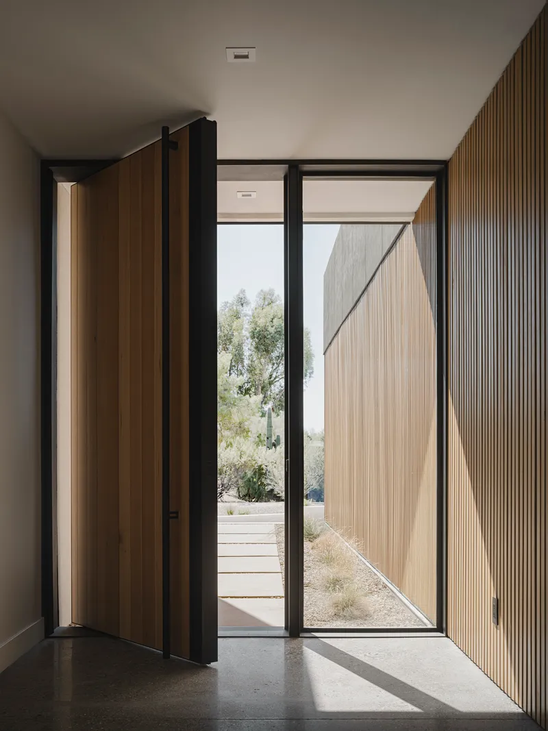 Entry vestibule with pivoting timber door and vertical fluted wood wall panel casting shadows on polished concrete floor