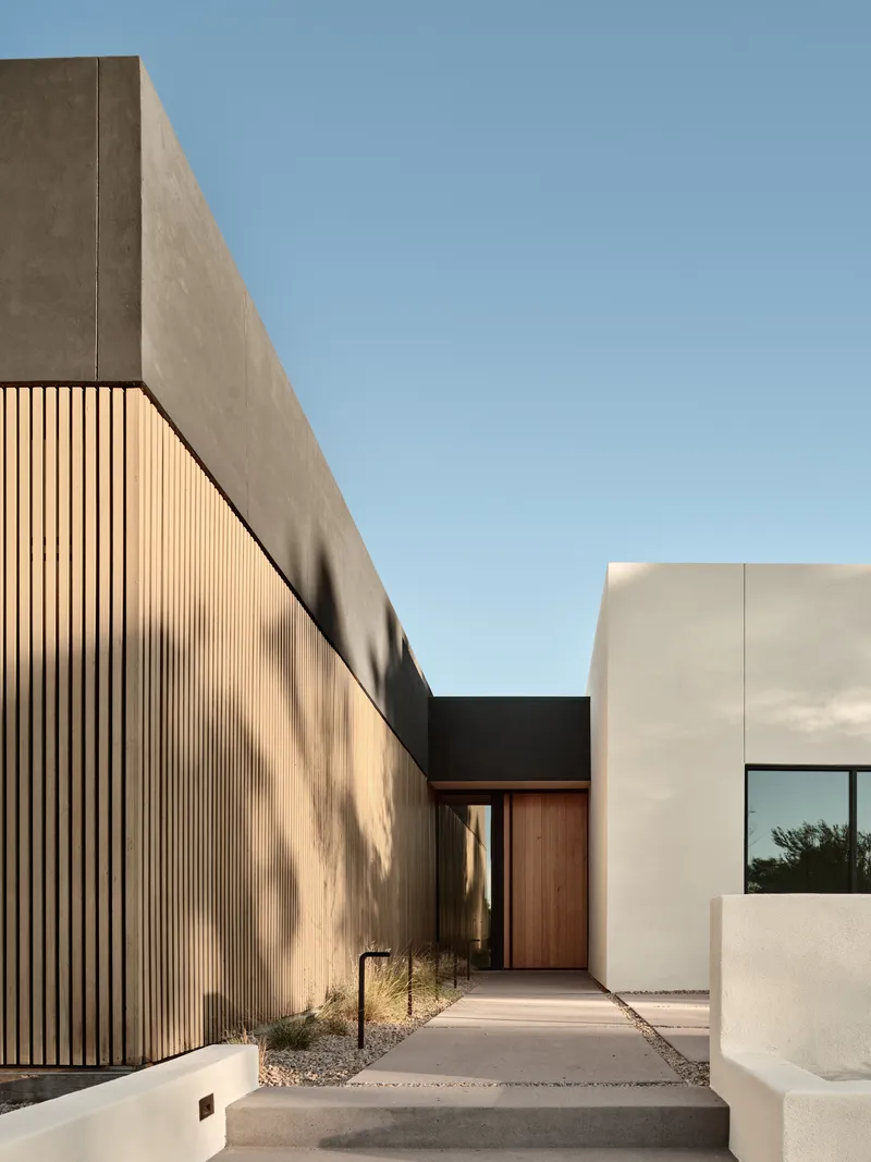 Entry walkway lined by vertical wood slat wall and concrete volumes casting afternoon shadows