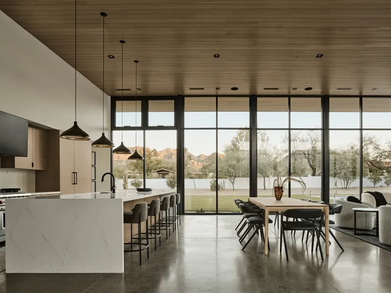 Open kitchen and dining space with floor-to-ceiling windows under wood plank ceiling at dusk