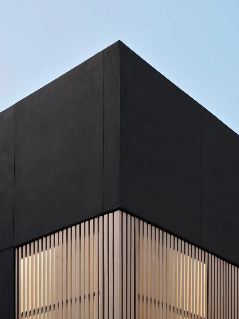 Corner detail of black cladding meeting illuminated timber slat screen at dusk
