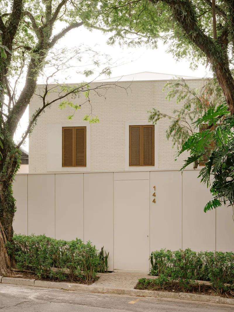 Front facade showing the white paneled wall with timber shutters and entrance door amid planted beds