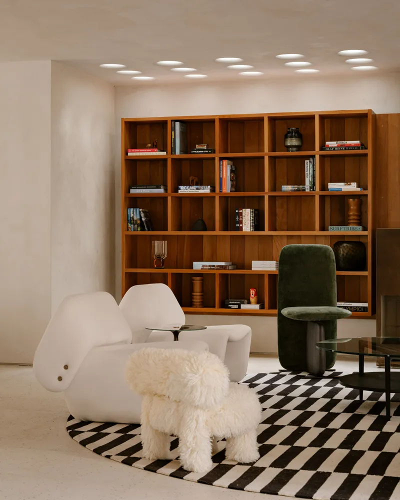 Living room with timber bookshelf wall and circular ceiling recesses illuminating a checkered rug