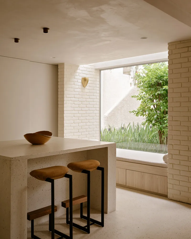 Kitchen counter with terrazzo surface and timber stools facing a large window opening to a planted courtyard