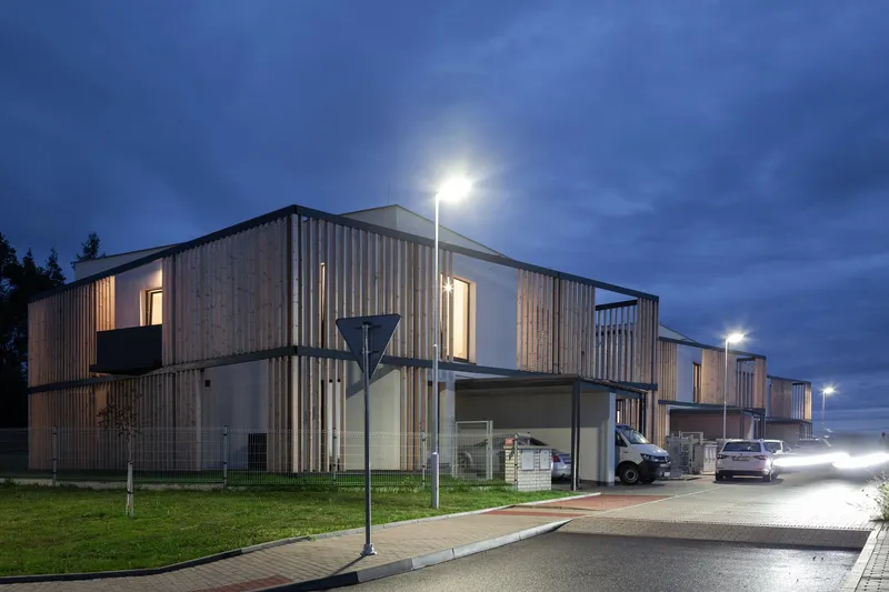 Evening view of the housing cluster, where timber screens glow softly, balancing privacy and openness within a cohesive urban edge.