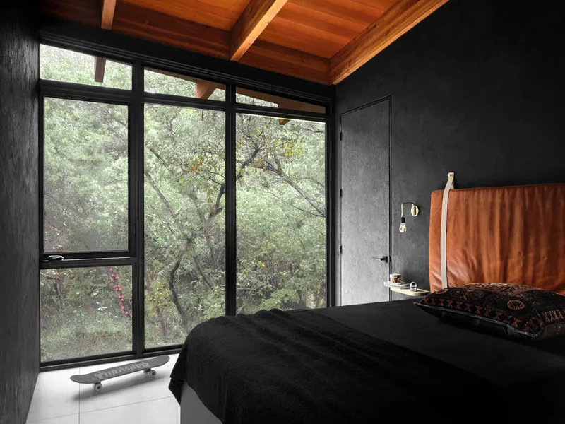 Bedroom with black-framed windows overlooking dense foliage and exposed timber ceiling beams