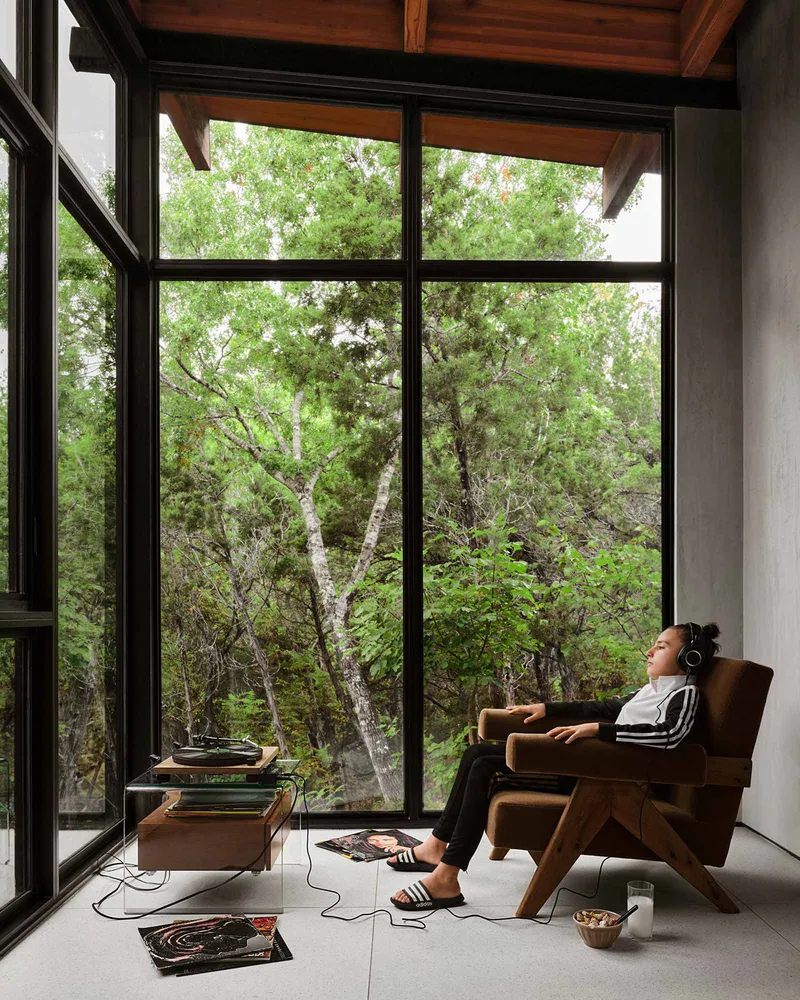 Interior seating nook with floor-to-ceiling windows framing trees where a person reclines listening to music