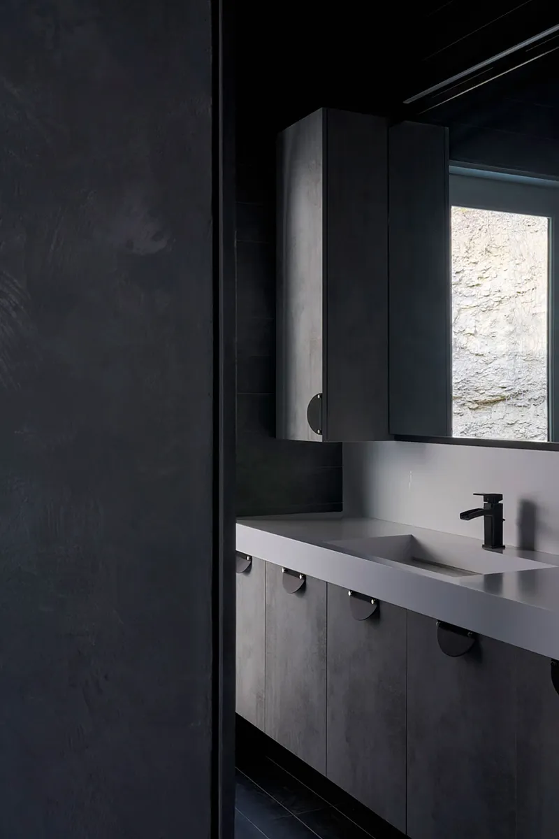 Bathroom vanity with concrete counter and narrow window revealing rough stone exterior wall