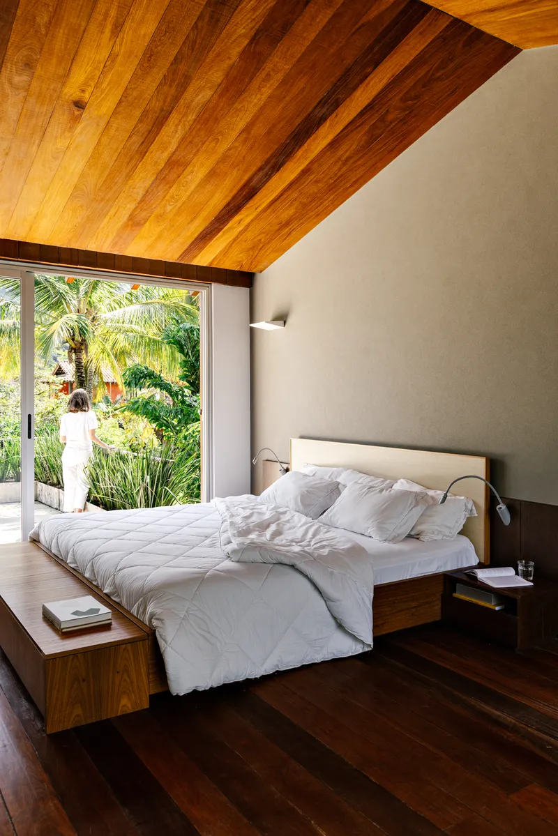 Bedroom with sloped timber ceiling and glazed wall opening to garden with palm trees