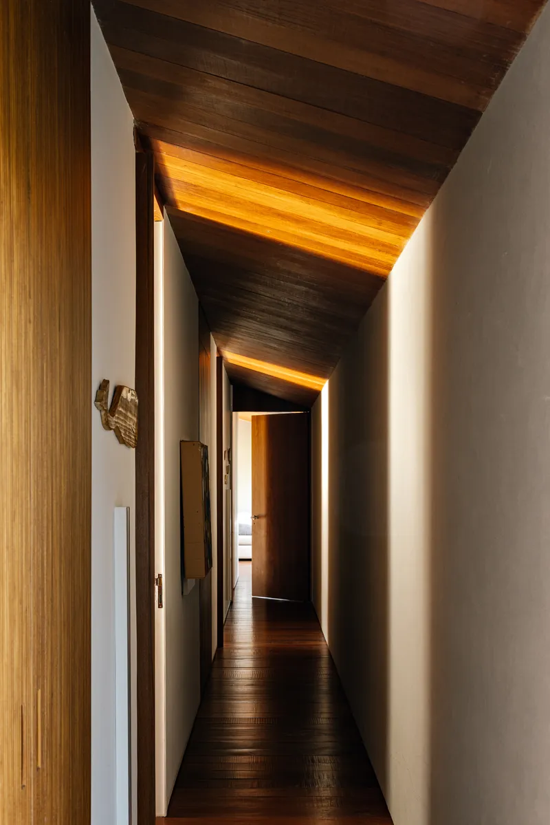Corridor with folded timber ceiling planes and concealed lighting washing the white walls at dusk
