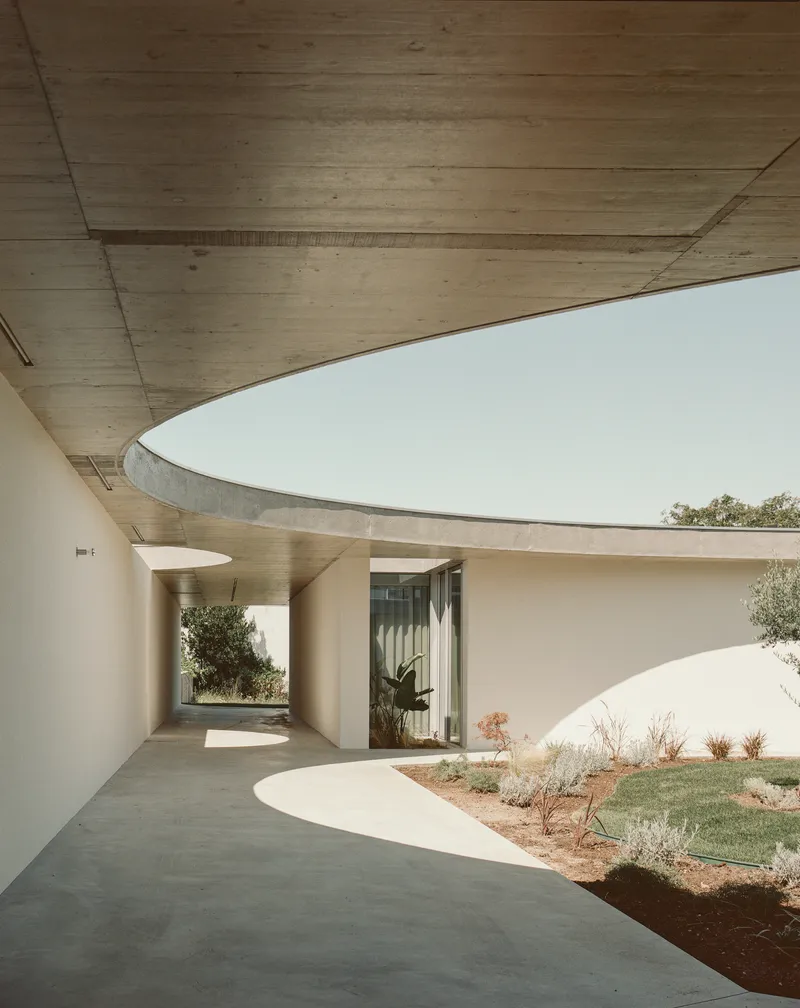 Curved covered walkway beneath exposed concrete ceiling with courtyard and plantings beyond