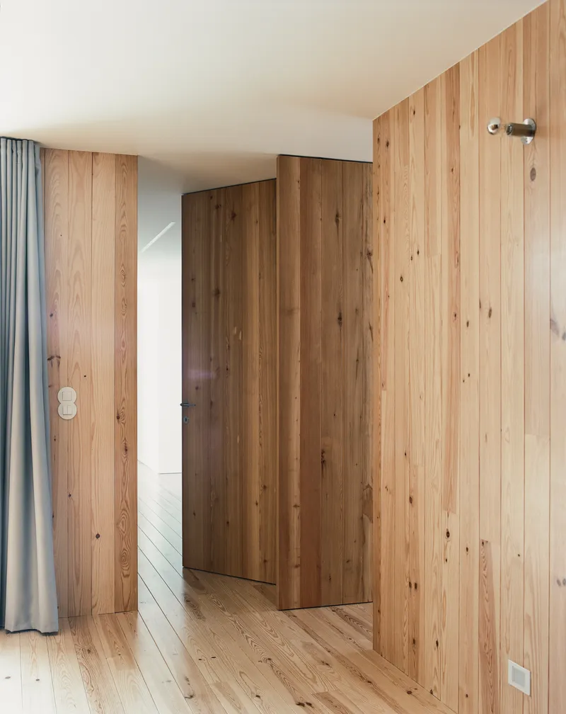 Vertical timber wall panels concealing a door beside a grey curtain under soft daylight