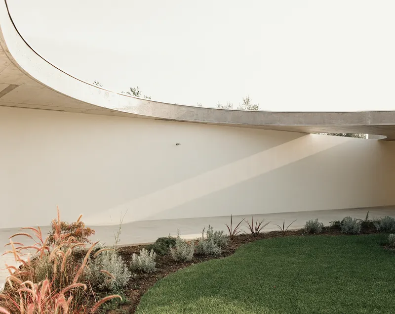 Concrete roof canopy curving over white walls with planted beds and lawn beneath