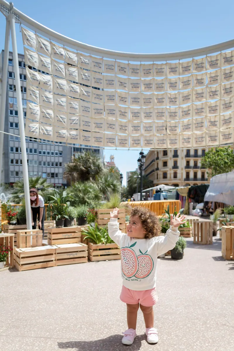 Child standing under the curved pavilion with timber furniture and planted beds in afternoon light