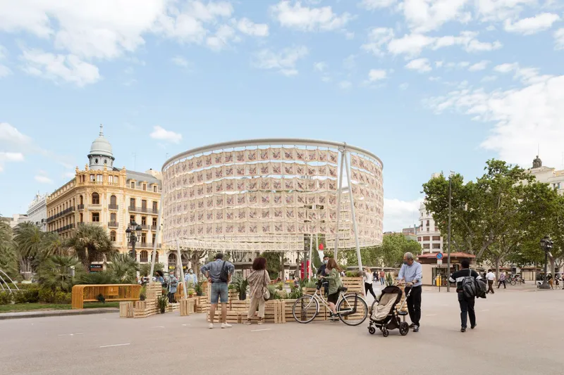 Cylindrical pavilion with patterned screen facade set in a public square with visitors and bicycles under sunny skies