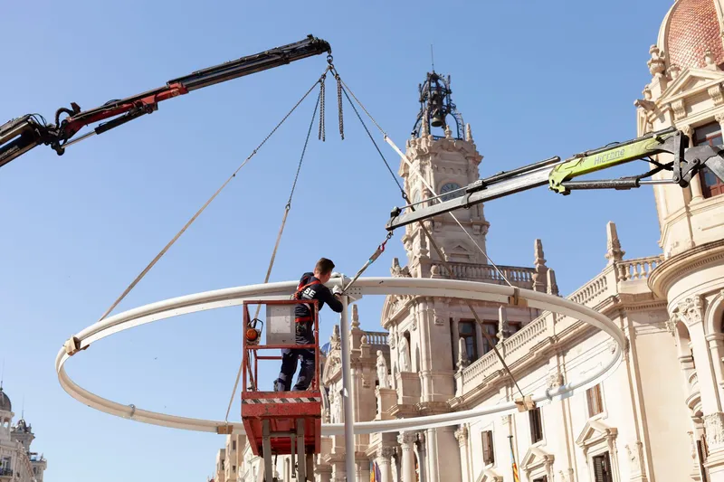 Worker on a lift installing a curved white structural ring suspended by cranes above surrounding historic facades