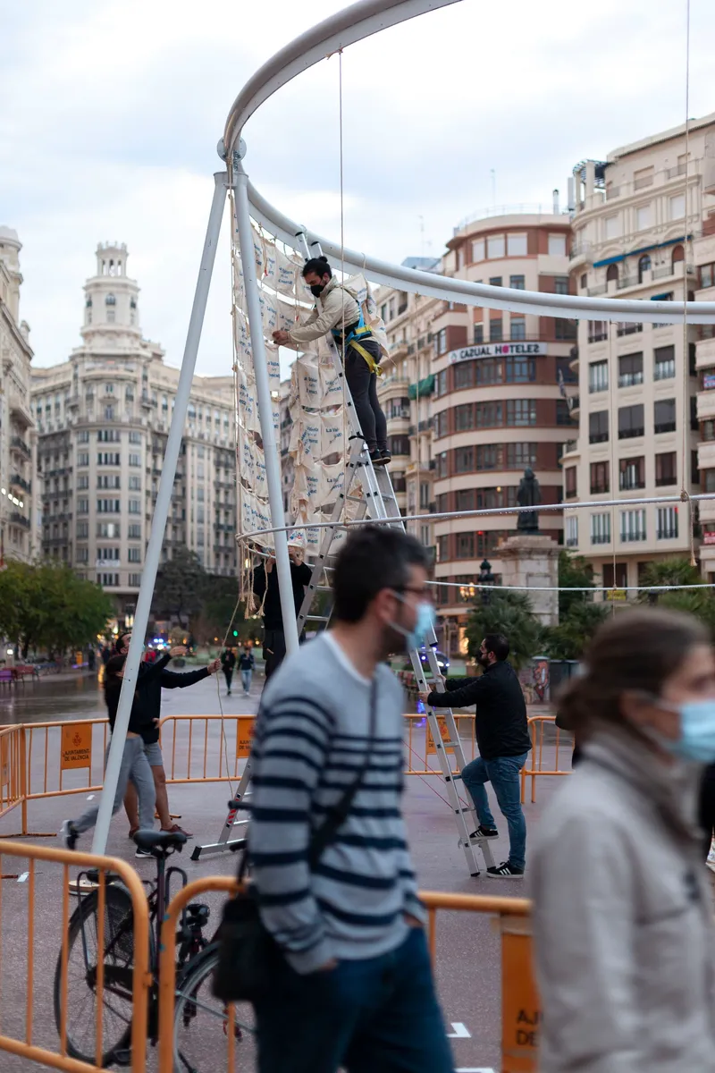 Temporary installation with curved metal frame and suspended fabric as pedestrians pass below wearing masks
