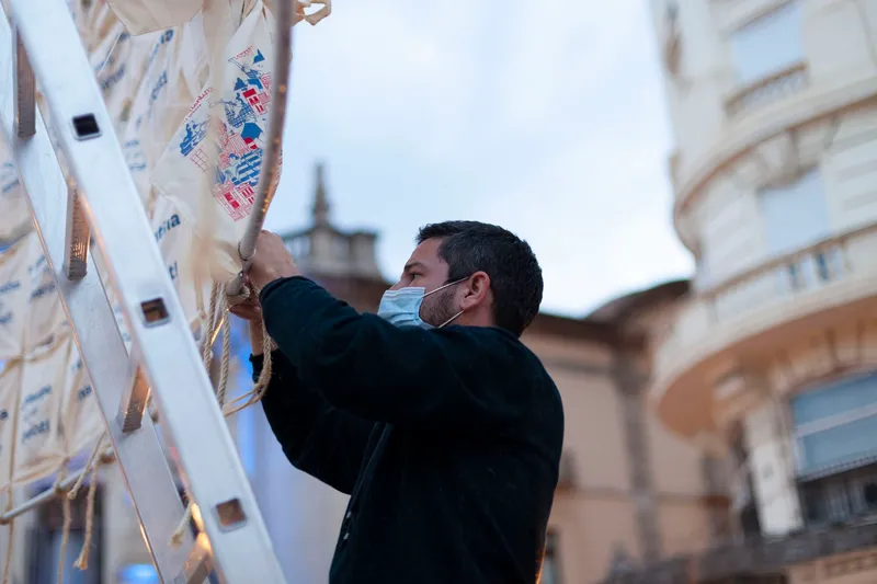Person in mask attaching printed textile panels to suspended rope against backdrop of building facades at dusk