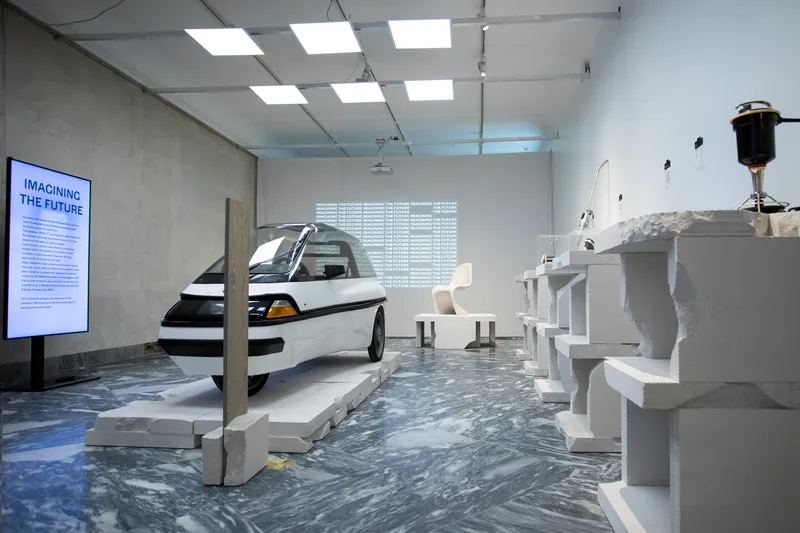 Gallery interior with concrete walls, suspended ceiling panels, and marbled grey flooring hosting sculptural objects