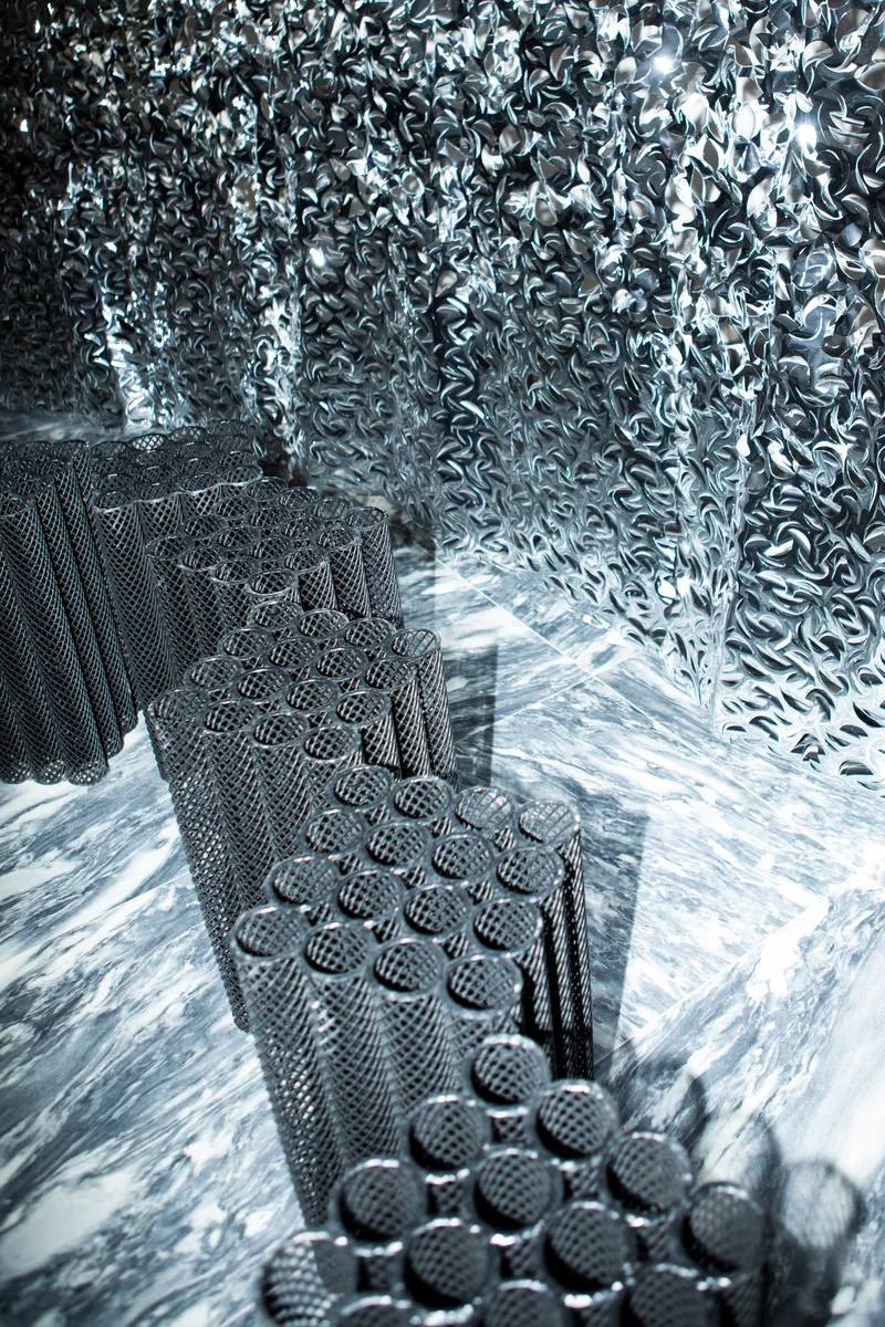 Textured black mesh staircase ascending through reflective metallic chain-link wall surface