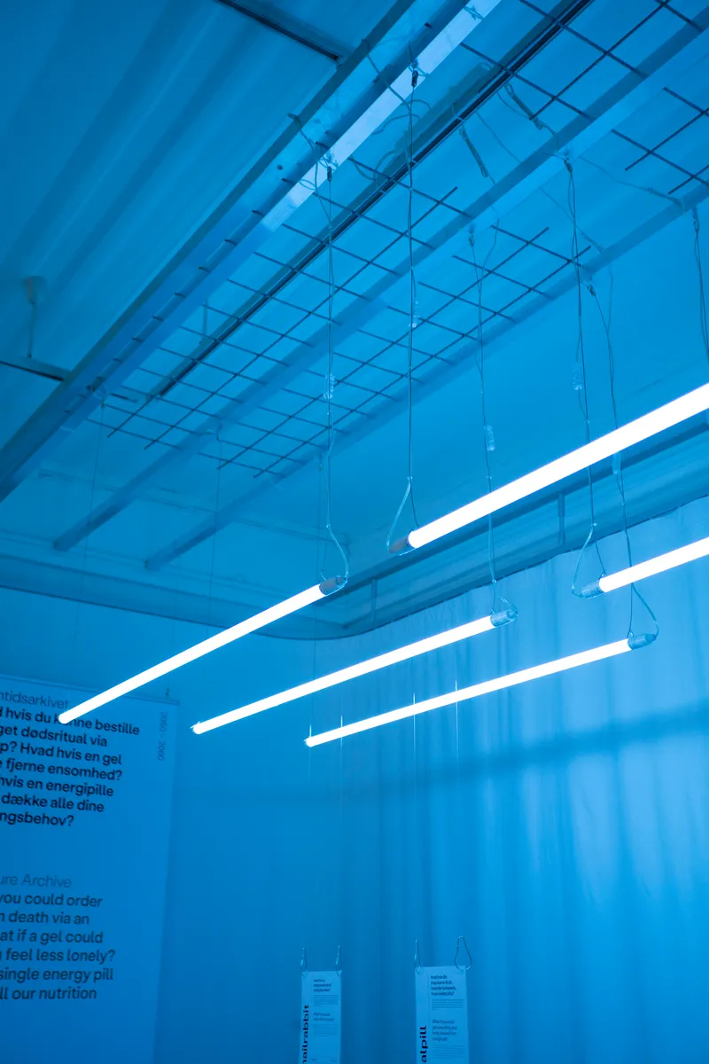 Exposed gridded ceiling with diagonal pendant light tubes and suspended information tags below