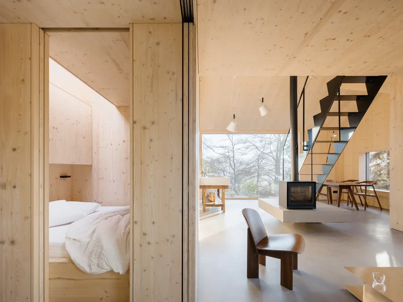 Open-plan interior showing bedroom alcove, cantilevered wood stove, and black steel staircase beside glazed wall