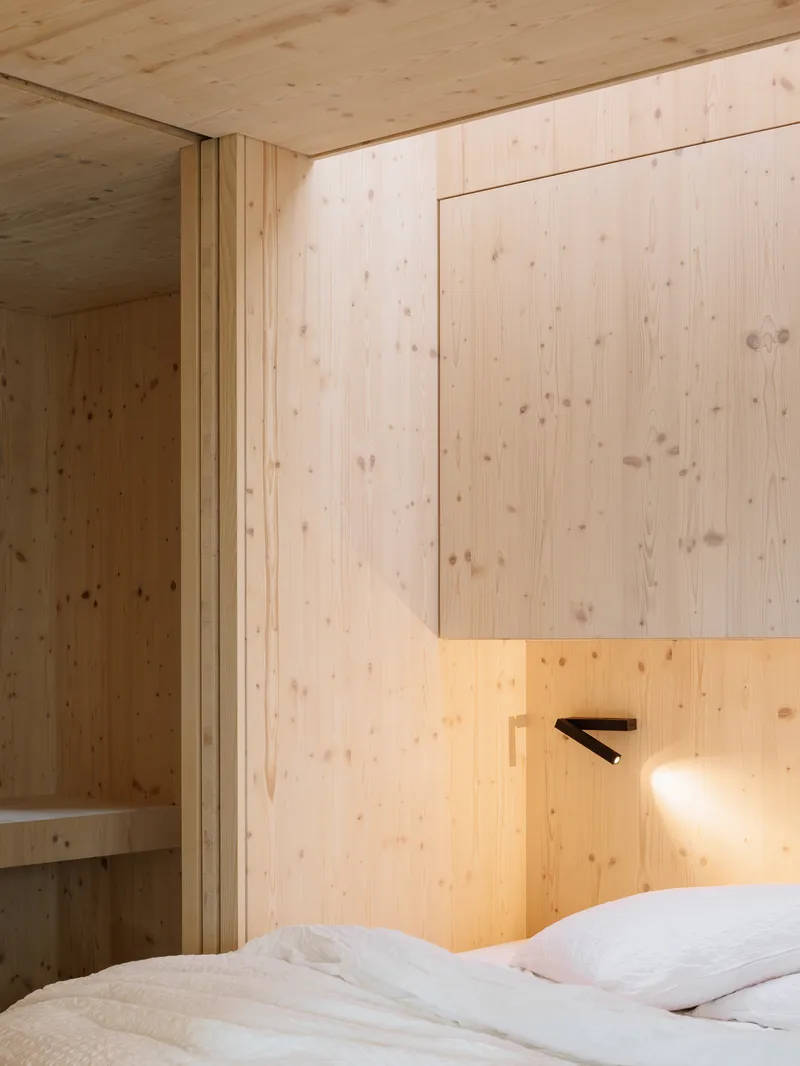 Bedroom corner with vertical timber paneling, recessed niche, wall-mounted reading light, and white bedding