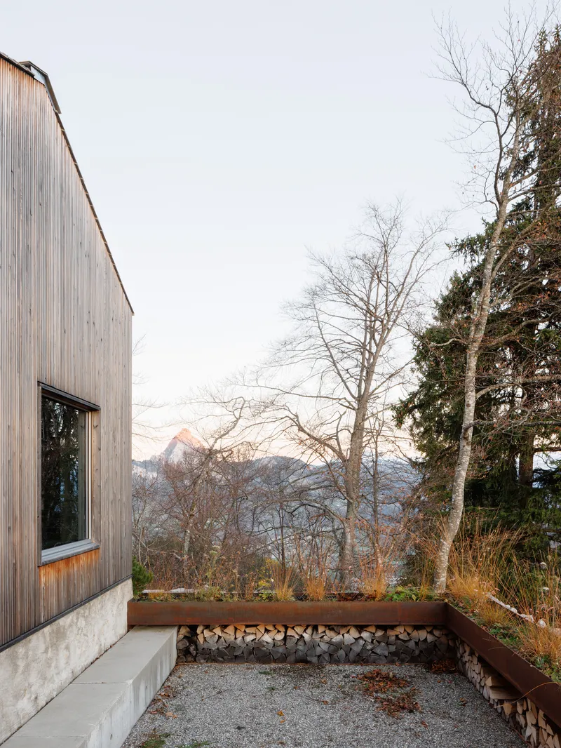 Gravel courtyard with concrete bench and corten steel planter beneath bare trees and distant peaks