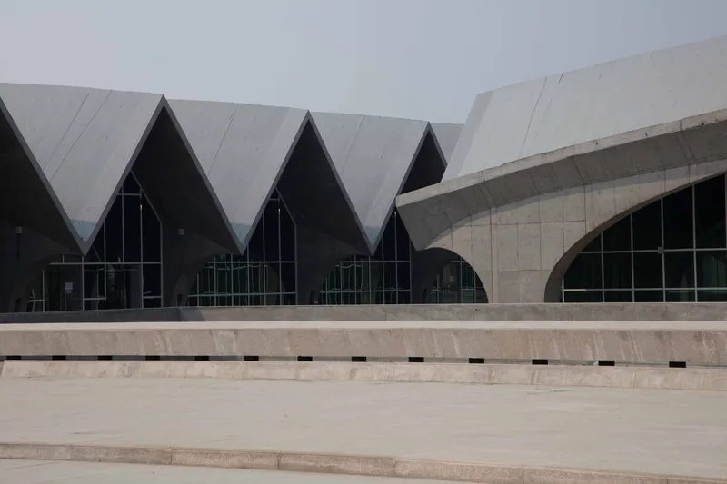 Concrete plaza with stepped seating facing a facade of zigzag gabled roofs and arched glass openings