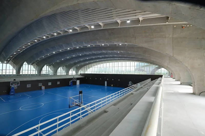 Concrete seating tiers overlooking a blue sports court beneath arched vaulted ceiling with linear skylights