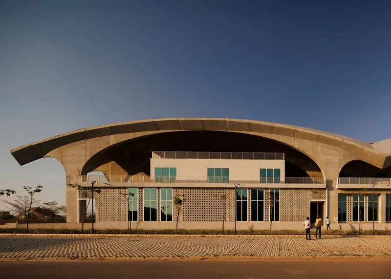 Curved concrete arches sheltering perforated brick facade with glazed openings at dusk with two figures on plaza